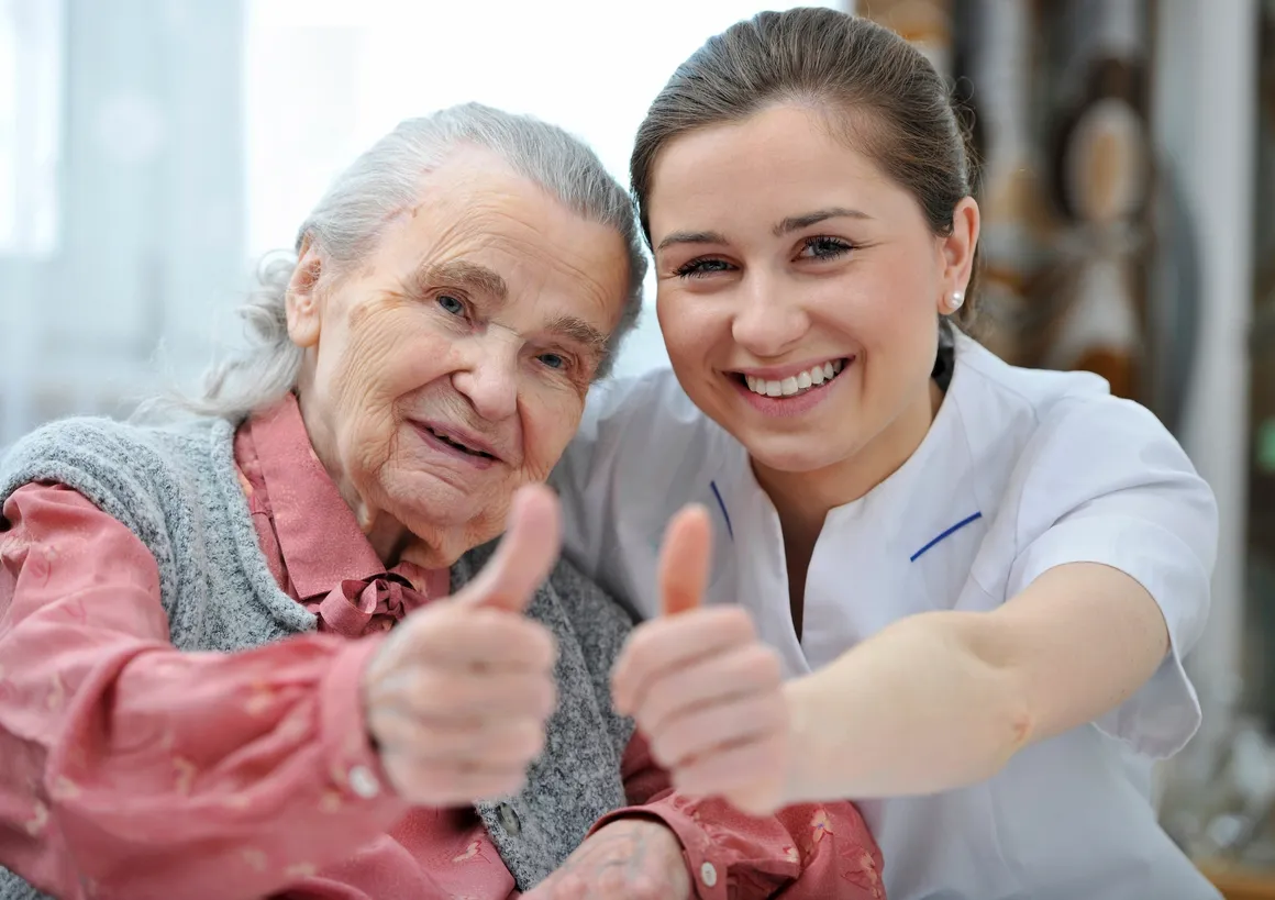 Elderly woman and caregiver facing the camera with thumbs up and smiles